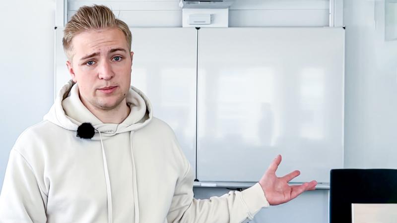 Auszubildender steht in einem weißen Hoodie vor einem Whiteboard. Er gestikuliert mit der linken Hand.
