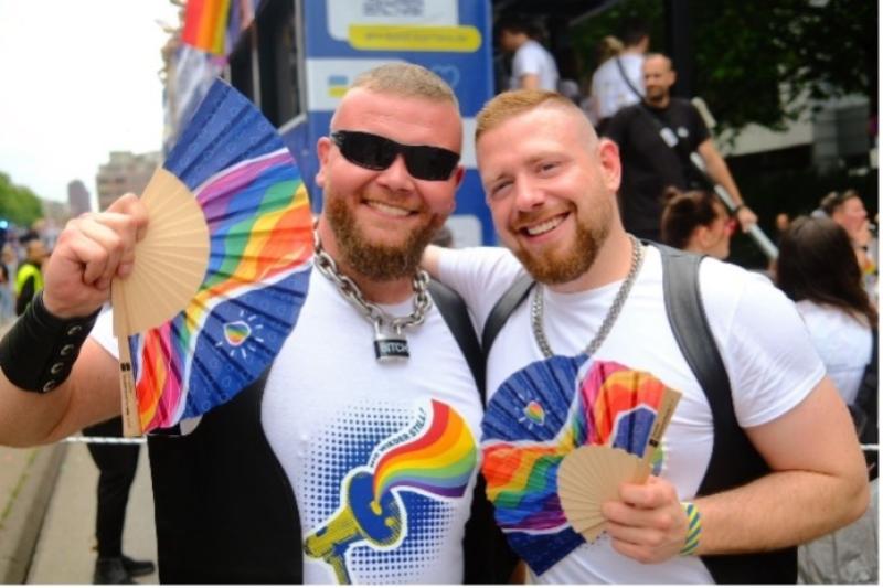 Zwei Personen mit Regenbogen-T-Shirts und Fächern bei einer Veranstaltung; im Hintergrund Menschen und ein blauer Bus.