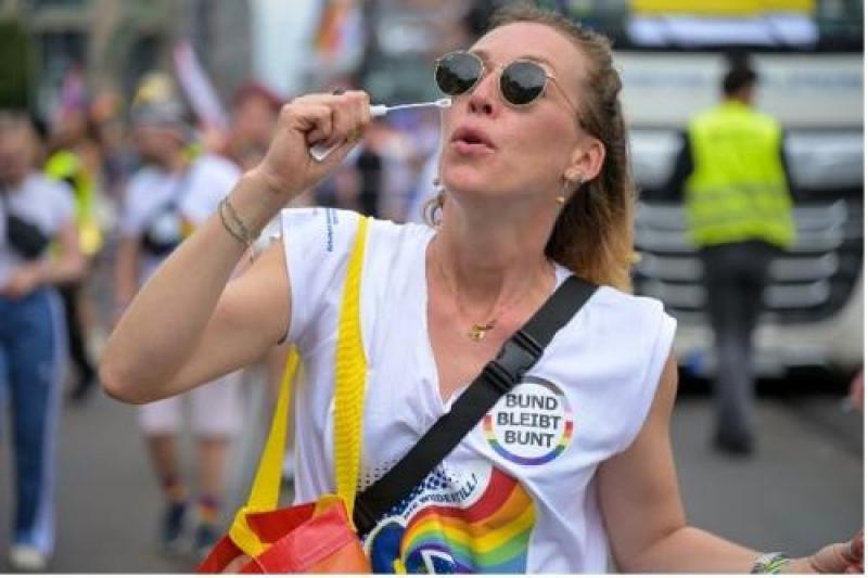 Person mit Regenbogen-Shirt und „BUND BLEIBT BUNT“-Badge bei öffentlicher Veranstaltung; hält Seifenblasenstab, im Hintergrund weitere Teilnehmende und ein Polizeifahrzeug.